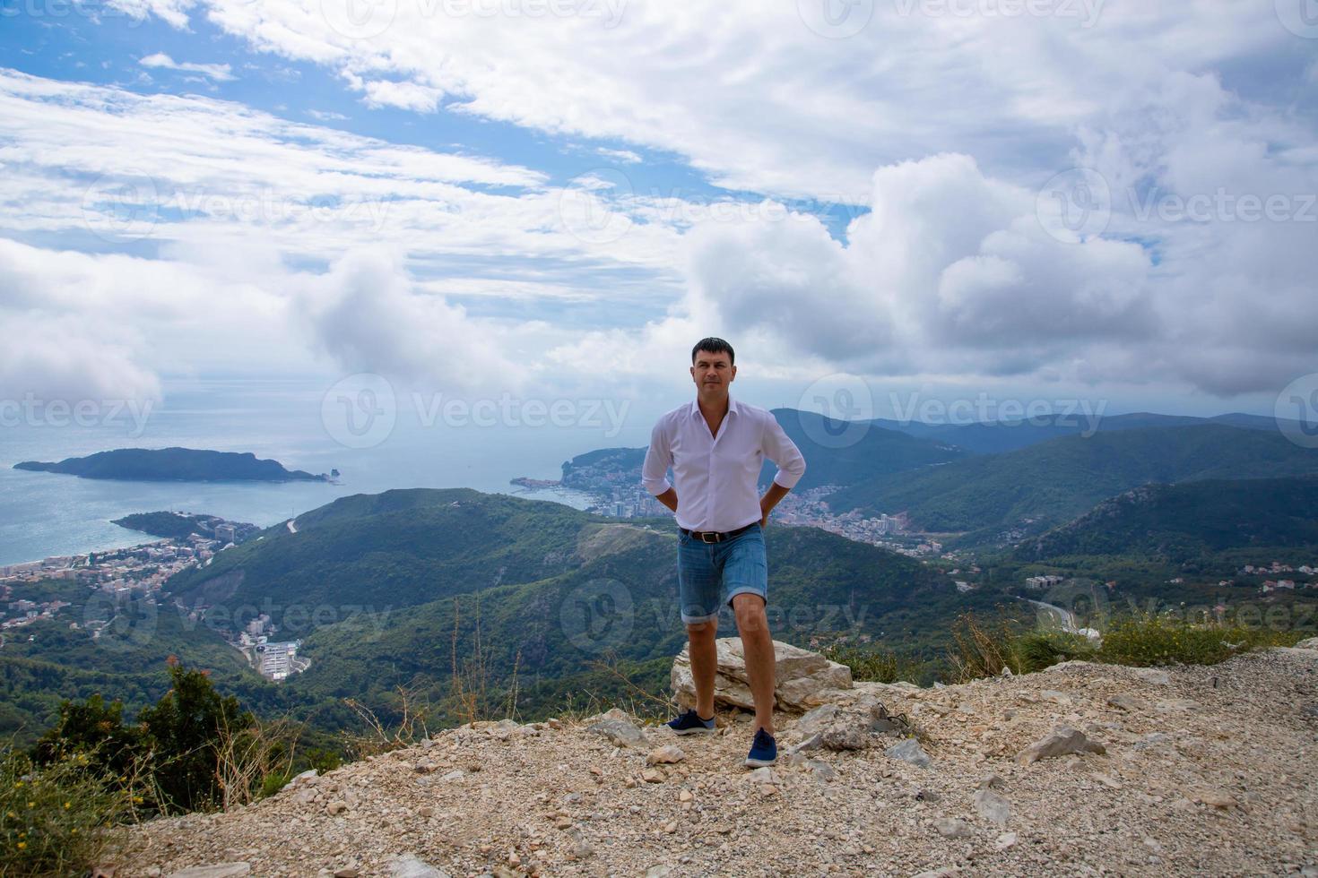 Caucasian man posing on the edge of a mountain peak with a gorgeous view of the Adriatic Sea and the city of Budva, Montenegro. photo