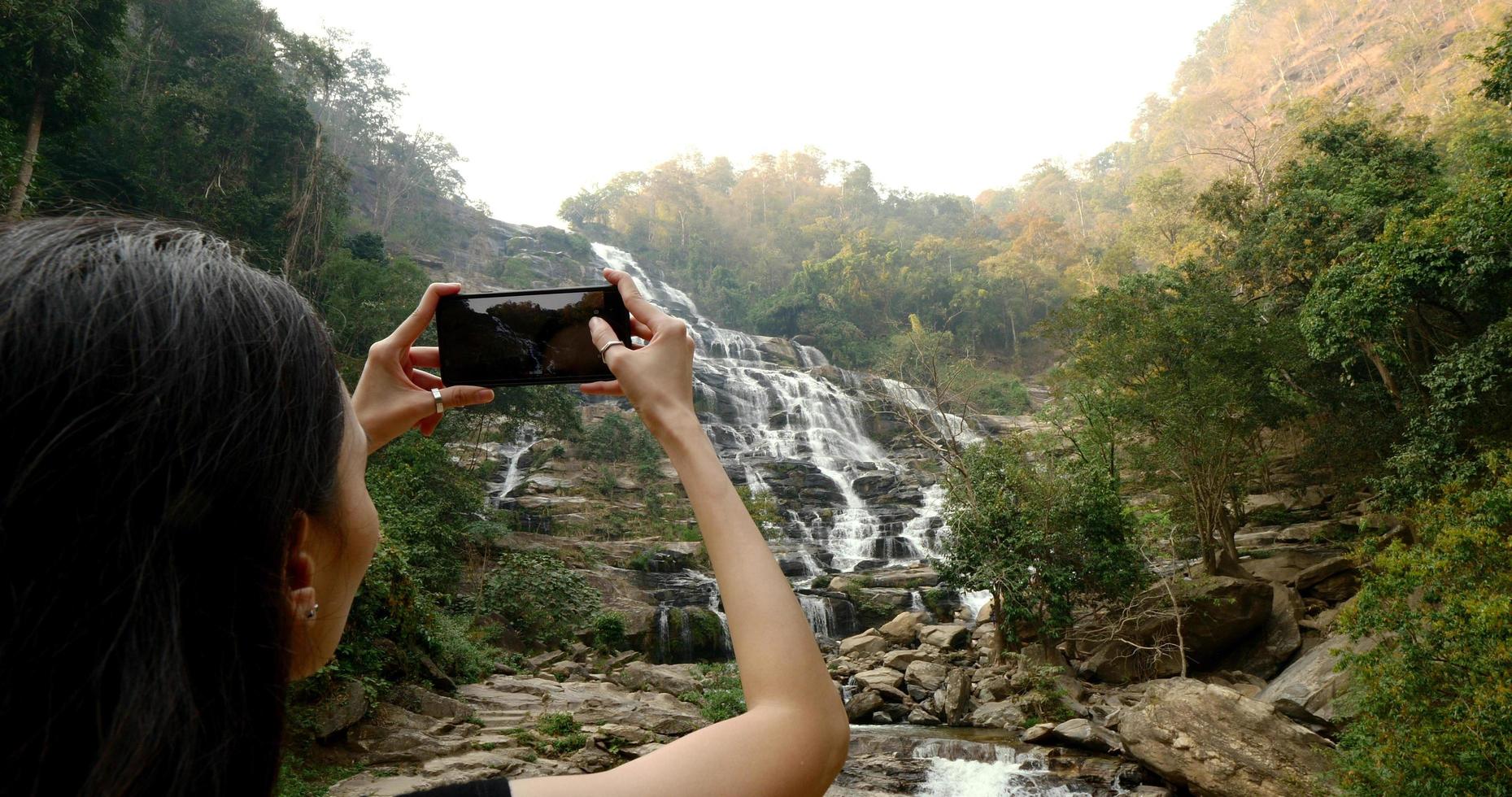 Woman in black t-shirt taking photos of beautiful waterfall by smartphone.