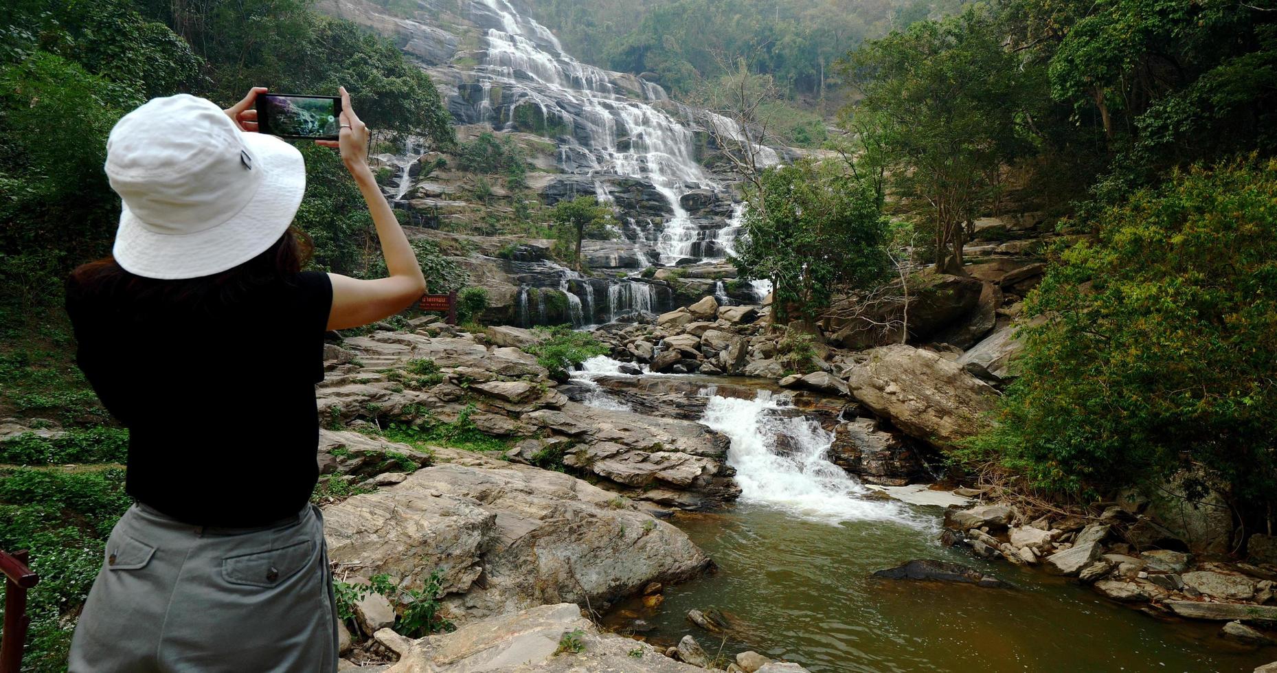 Woman in black t-shirt taking photos of beautiful waterfall by smartphone.
