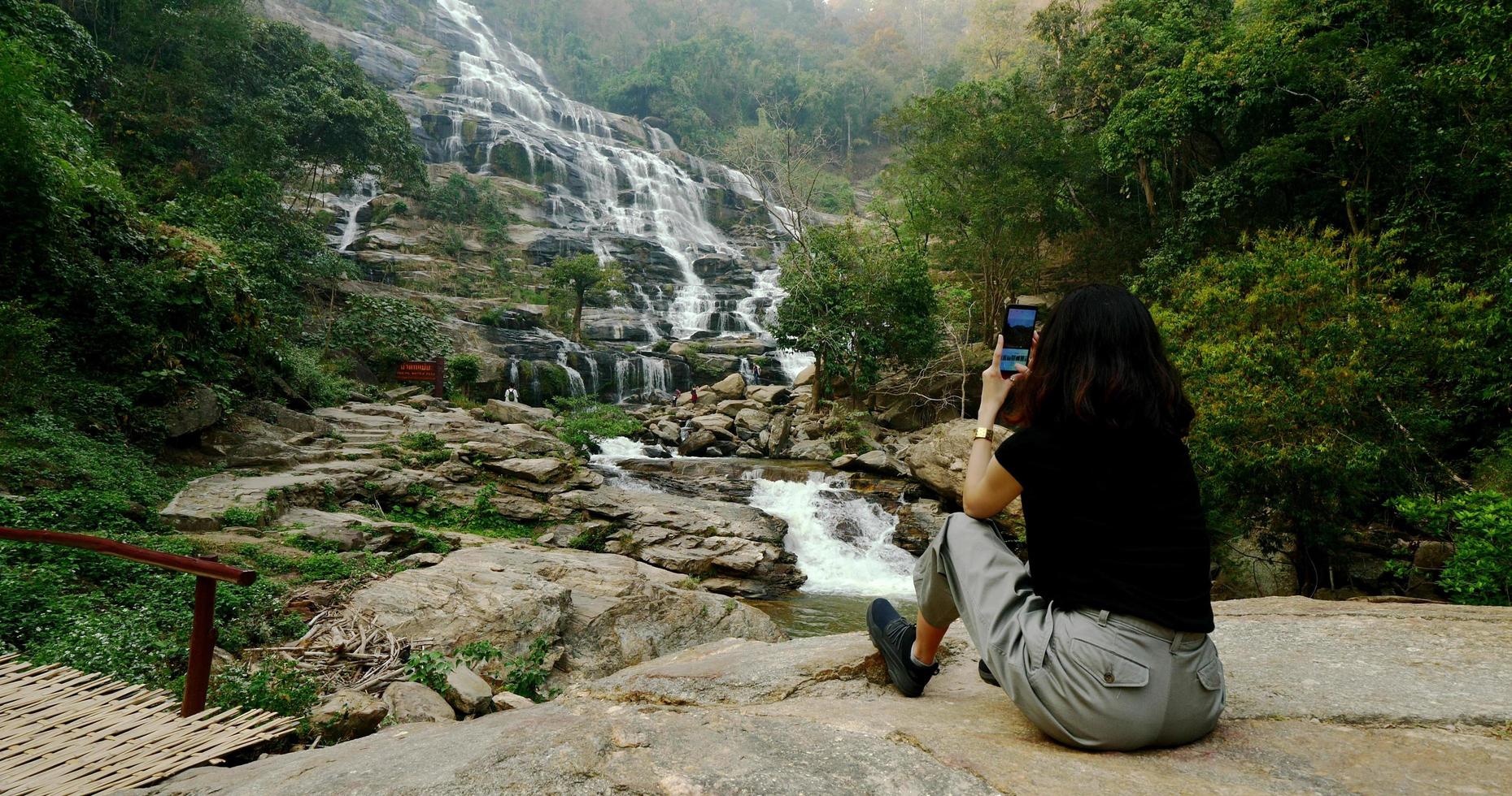 Woman in black t-shirt sitting and taking photos of beautiful waterfall by smartphone.