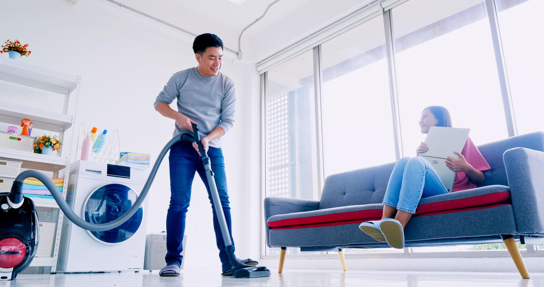 el marido limpia el suelo con una aspiradora mientras la mujer trabaja con un portátil en el sofá. feliz pareja asiática pasando tiempo juntos en casa. foto
