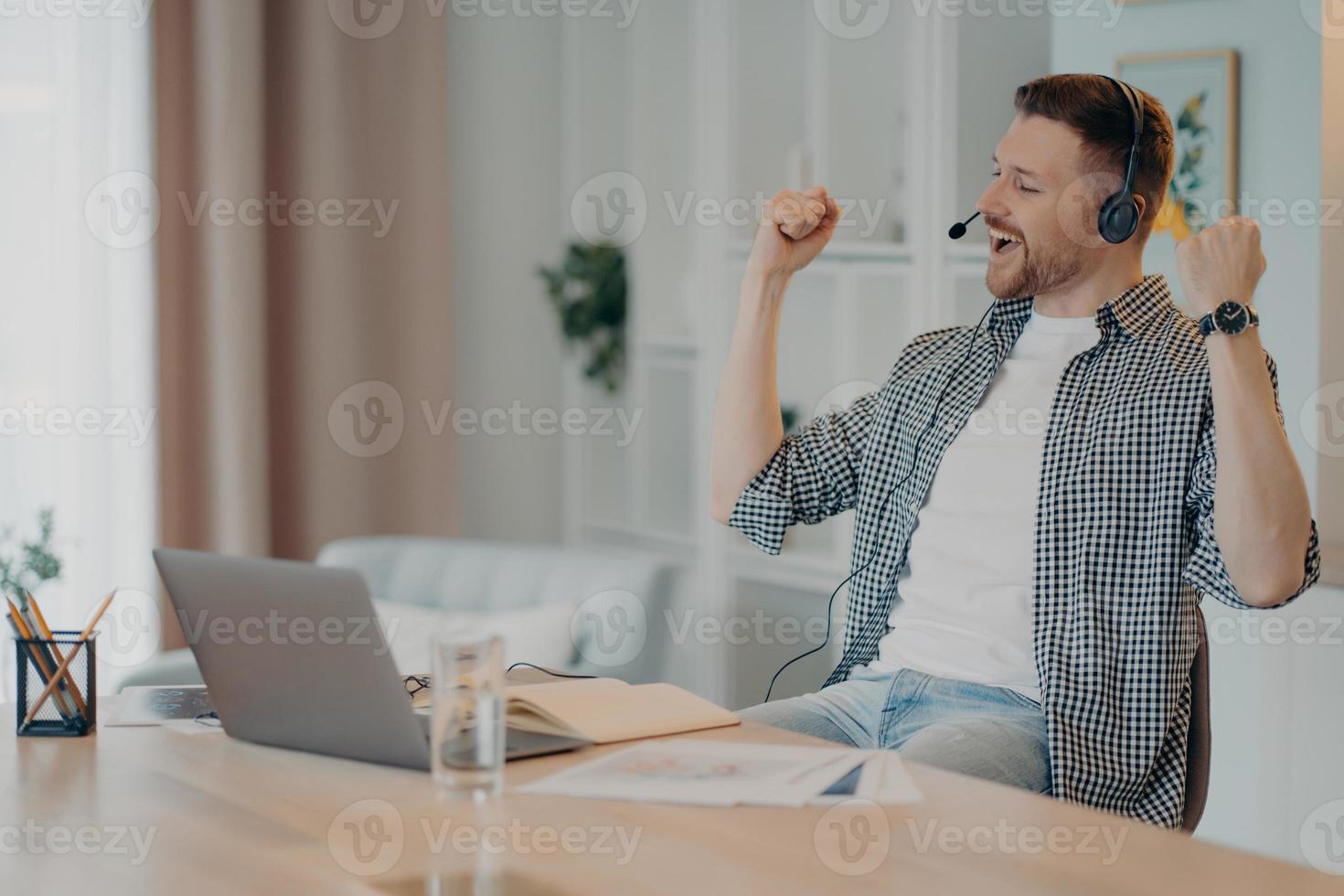 Happy successful man clenches fists celebrates triumph works online wears headset poses at desktop with laptop computer dressed casually. Overjoyed male student listens educational online lecture photo