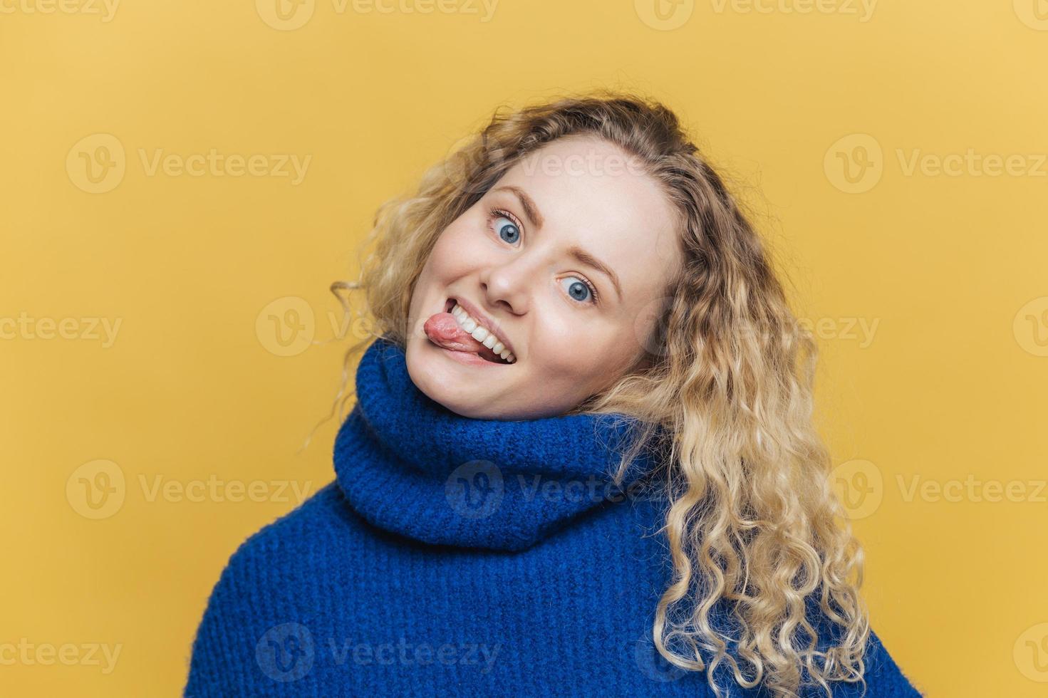 Funny comic curly young woman with positive expression, shows tongue, has fun with friends, wears blue sweater, makes grimace, isolated over yellow studio background. People and fun concept. photo