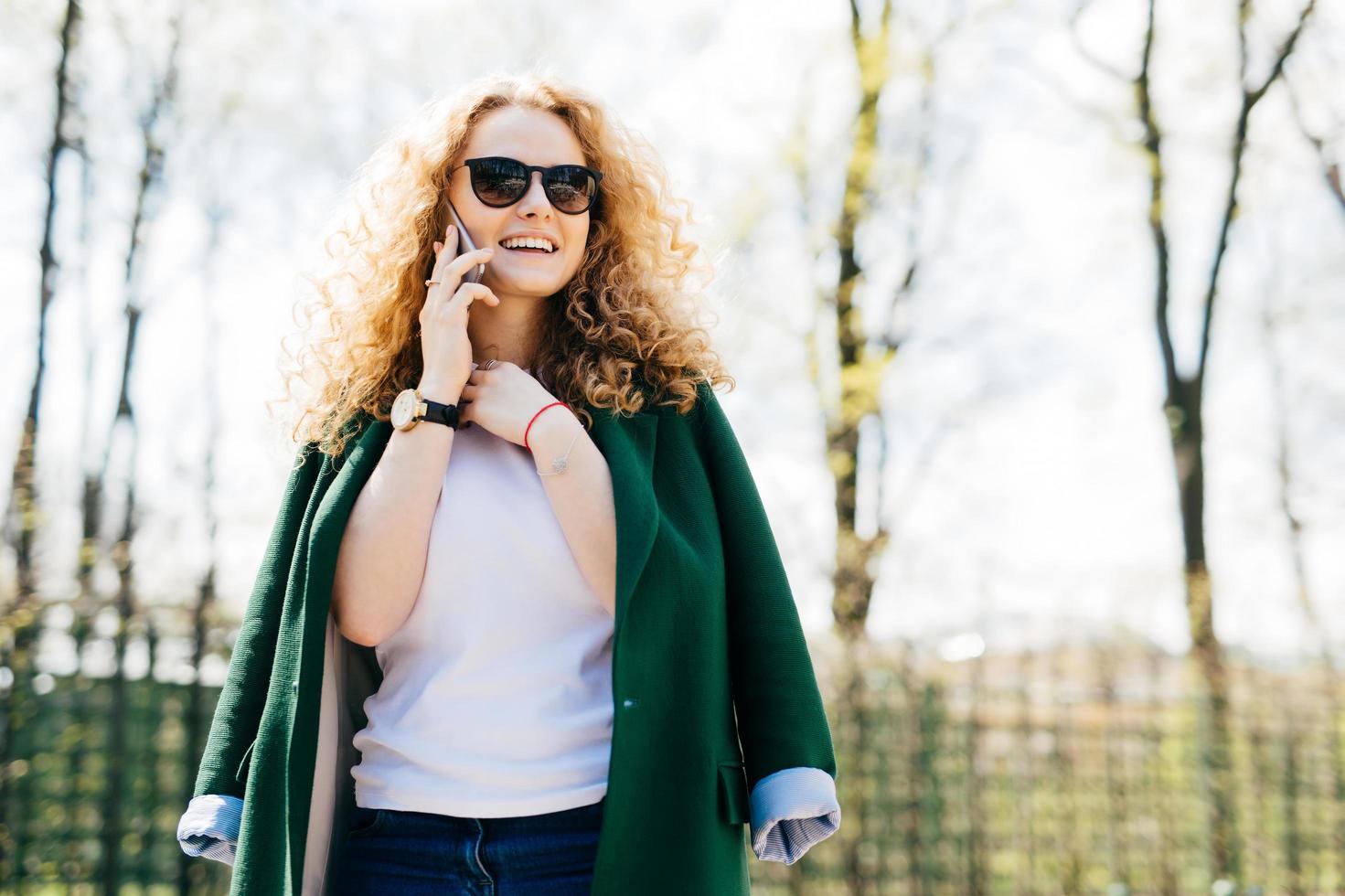 Portrait of beautiful female with fluffy blonde hair talking on cell phone to her friend, looking happily and excited standing against green park background holding hand on neck. Human emotions photo