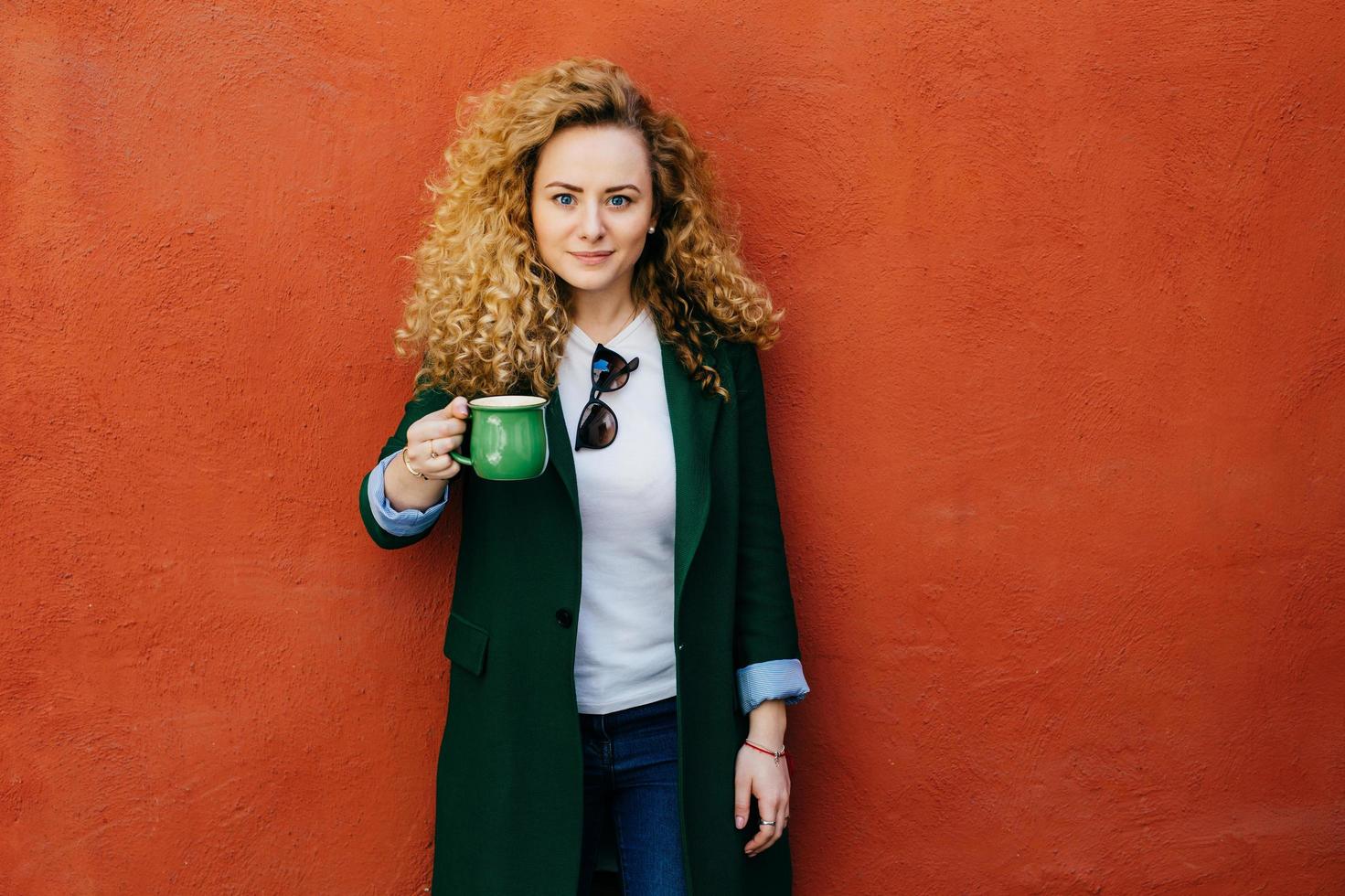 Beautiful confident blonde curly woman wearing elegant jacket and jeans holding green mug with tea posing against orange background in studio. People, free time, lifestyle and relaxation concept photo