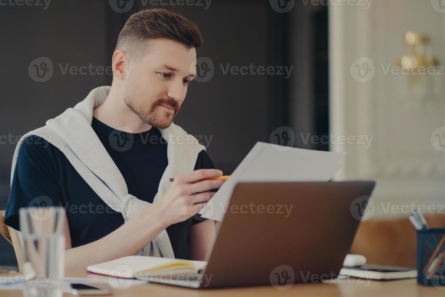 Concentrated man reads information or data from paper documents works on freelance poses at desktop against cozy interior dressed in casual clothes busy working. Male entrepreneur at coworking space photo