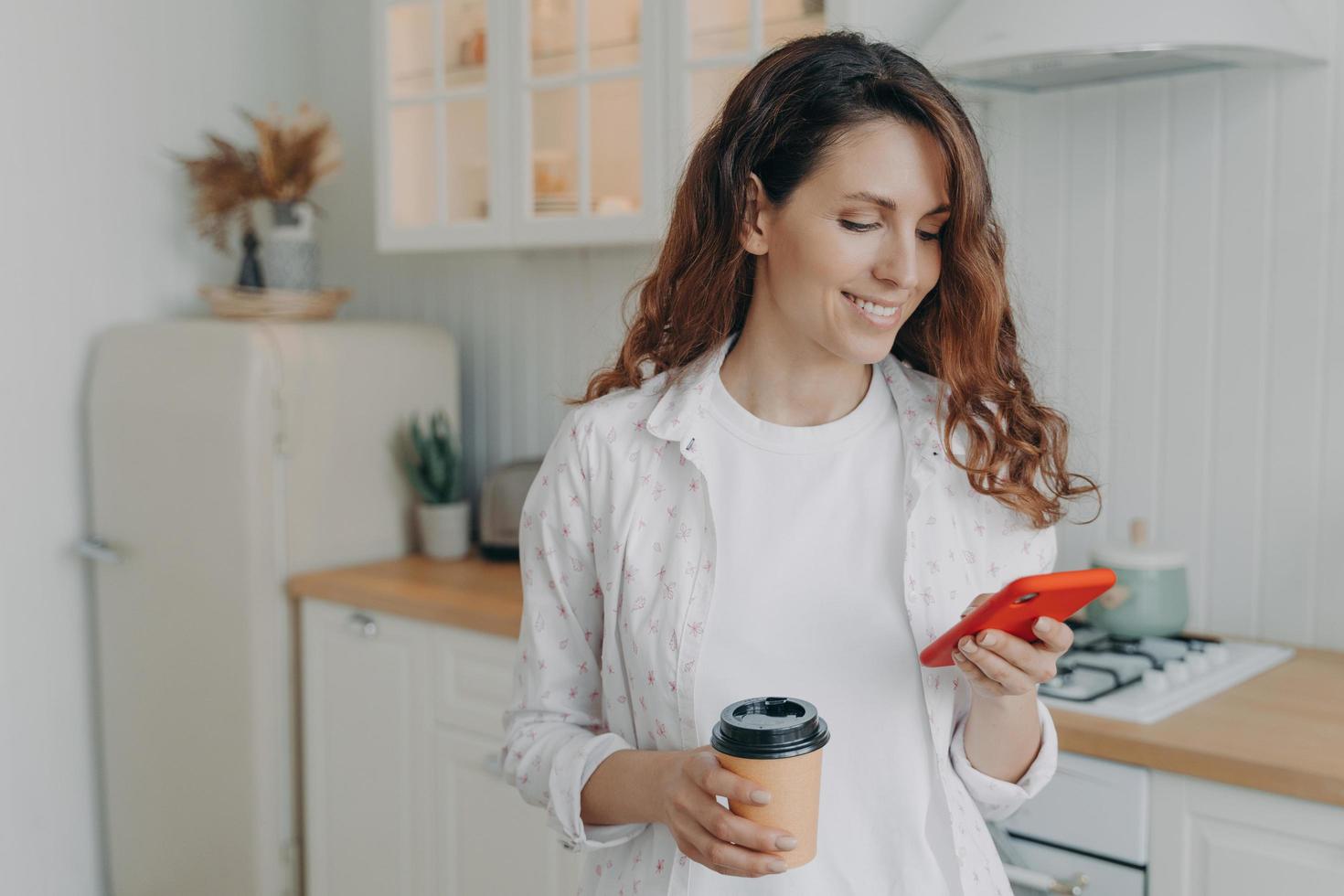 European woman with smartphone has coffee. Girl is reading message in kitchen at home and smiling. photo