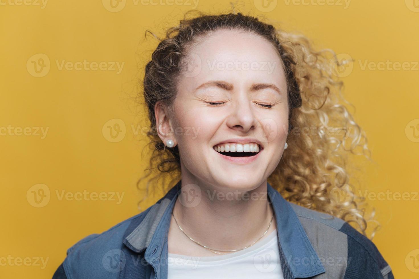 Overjoyed beautiful female laughs joyfully as hears good joke, smiles broadly, keeps eyes shut, has curly hair, isolated over yellow background. Cute young woman poses indoor, expresses happiness photo