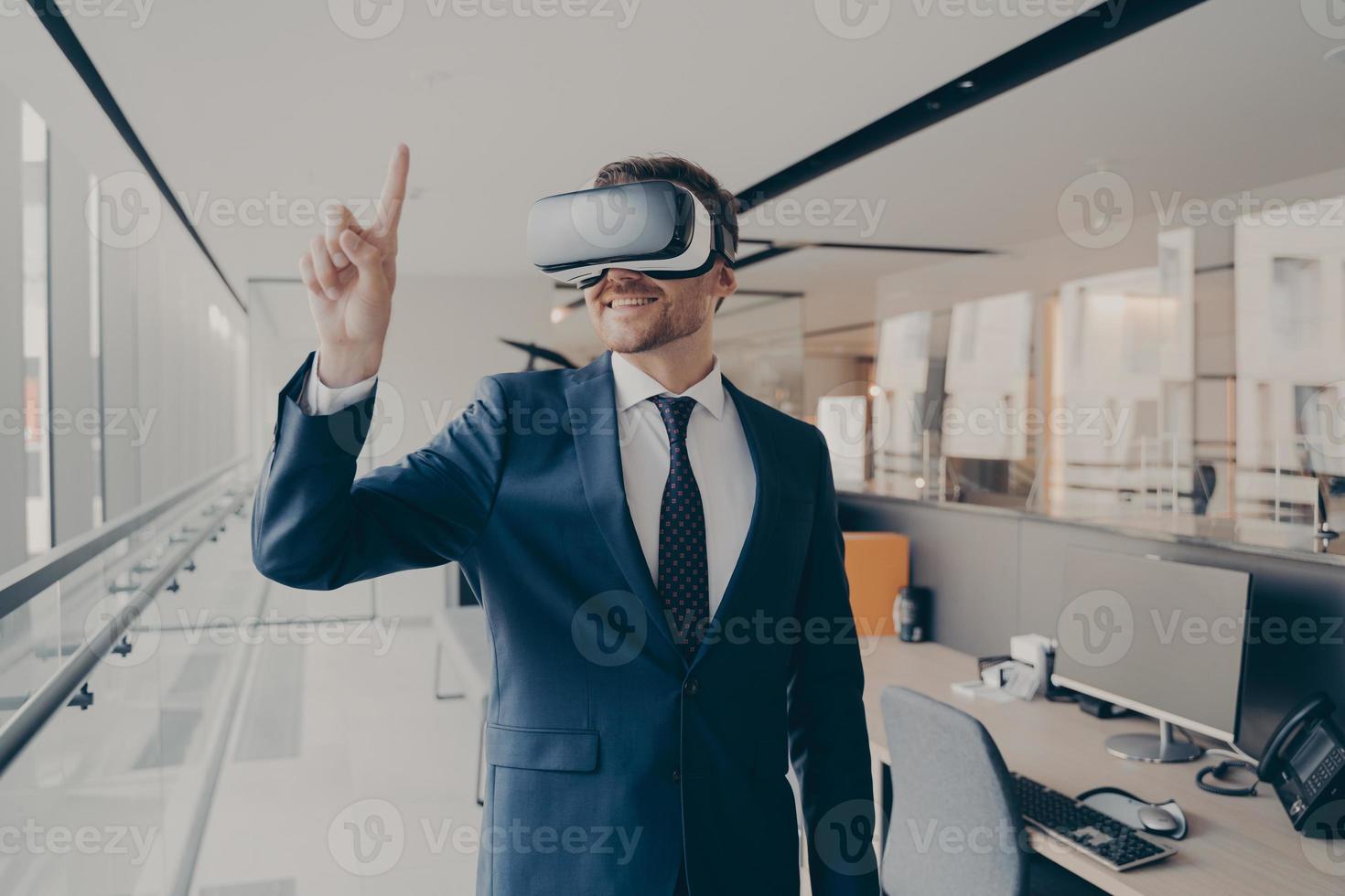 Happy man office worker wearing vr goggles, pointing with finger at objects in digital world photo