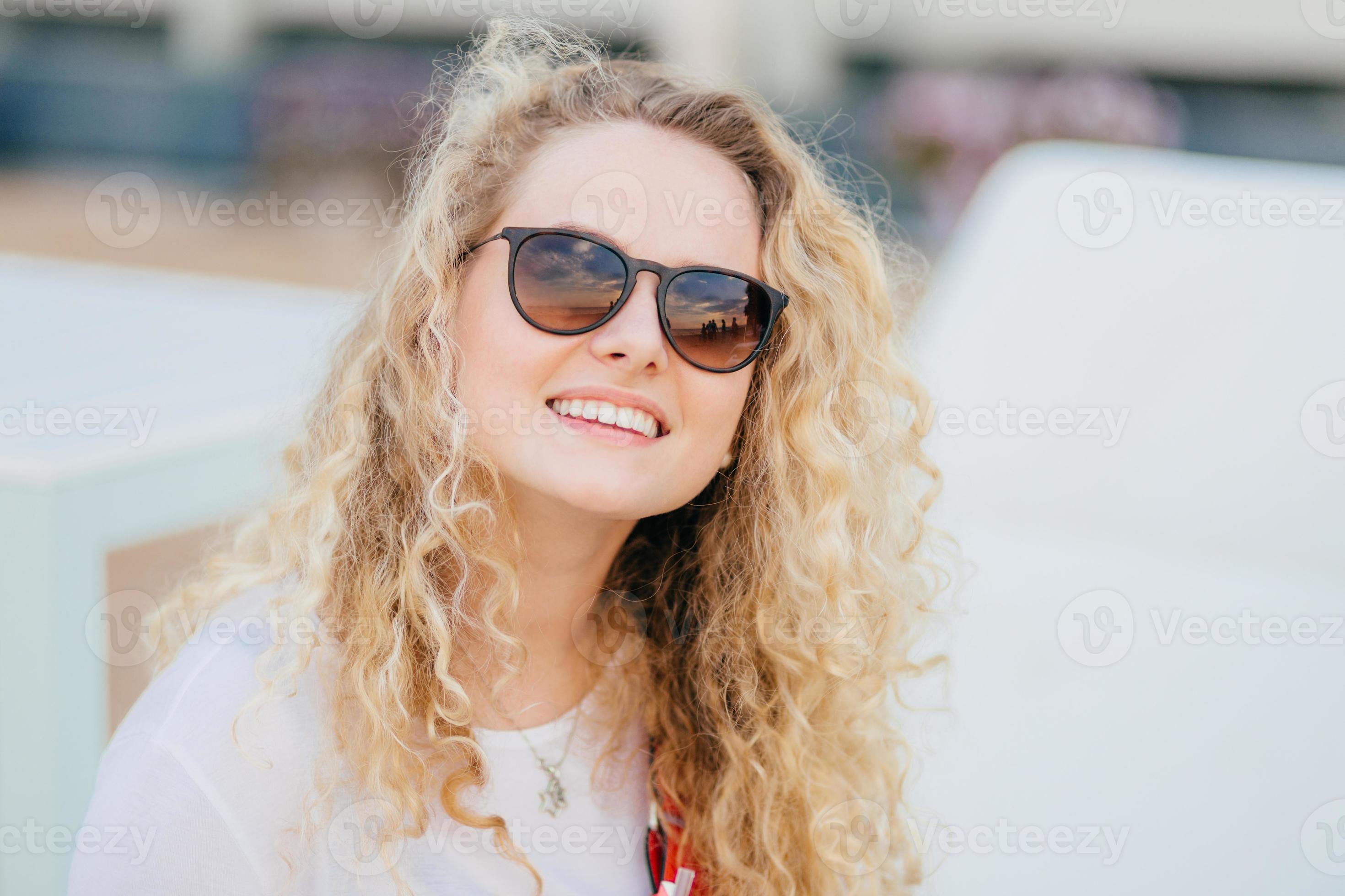 Photo of attractive young female has light curly hair, wears trendy shades,  has toothy smile, shows white teeth, recreats outdoor, smiles as recieves  compliment from boyfriend. Summer time, rest 11099849 Stock Photo