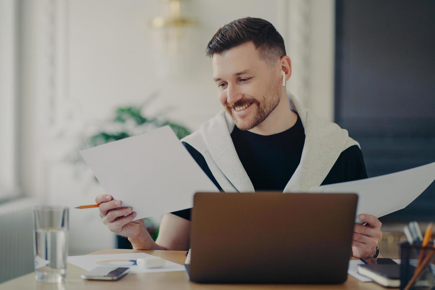 Handsome happy businessman analyzing documents and feeling satisfied while working on laptop from home photo