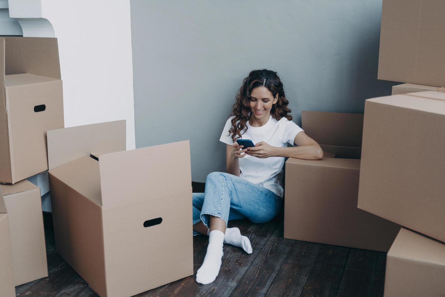 Girl texting on mobile phone and sitting on the floor surrounded with cardboard packages and boxes. photo