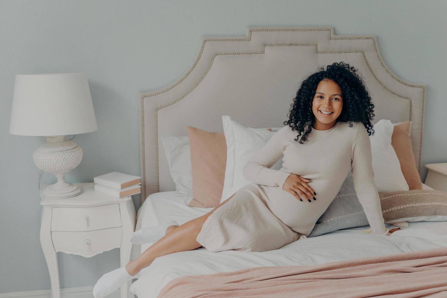 Happy pregnant african american lady resting on bed in modern apartment photo