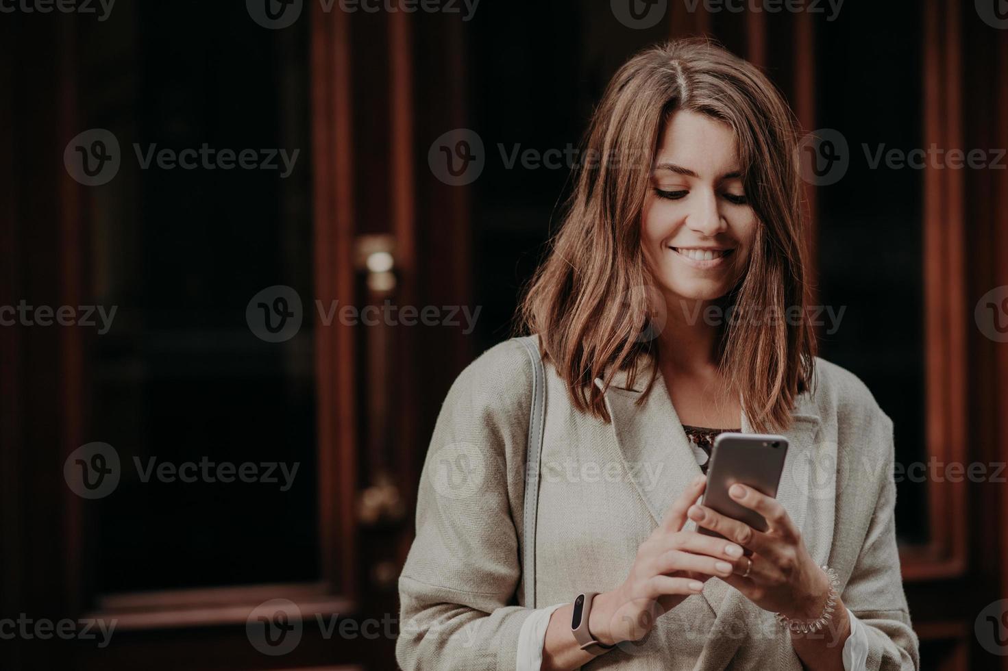 Horizontal shot of pretty cute brunette female model shares multimedia files via cell phone, dressed in elegant clothes, reads positive news in internet website, connected to wireless internet. photo
