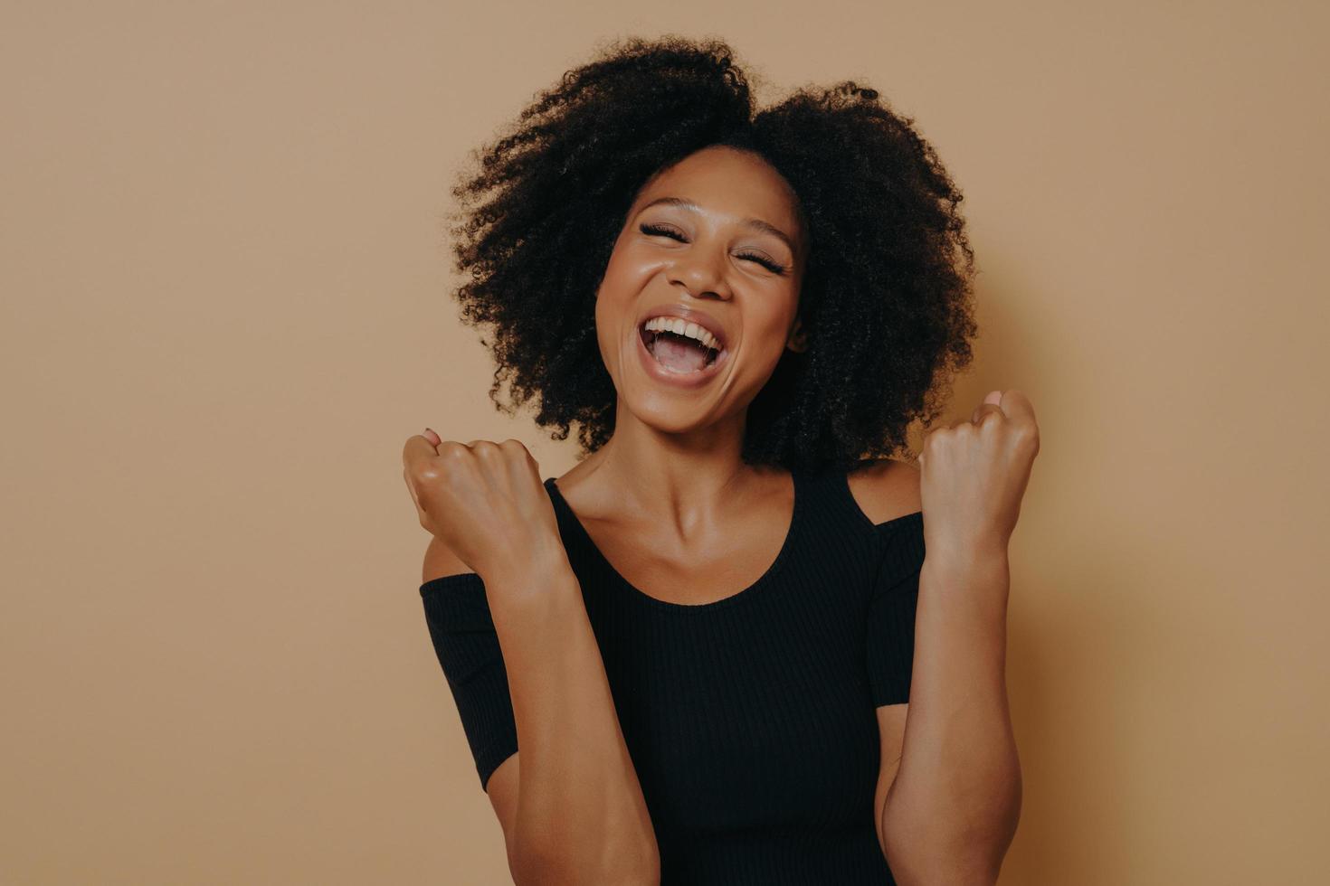 Young joyful excited dark skinned woman keeping fists clenched while posing against beige background photo