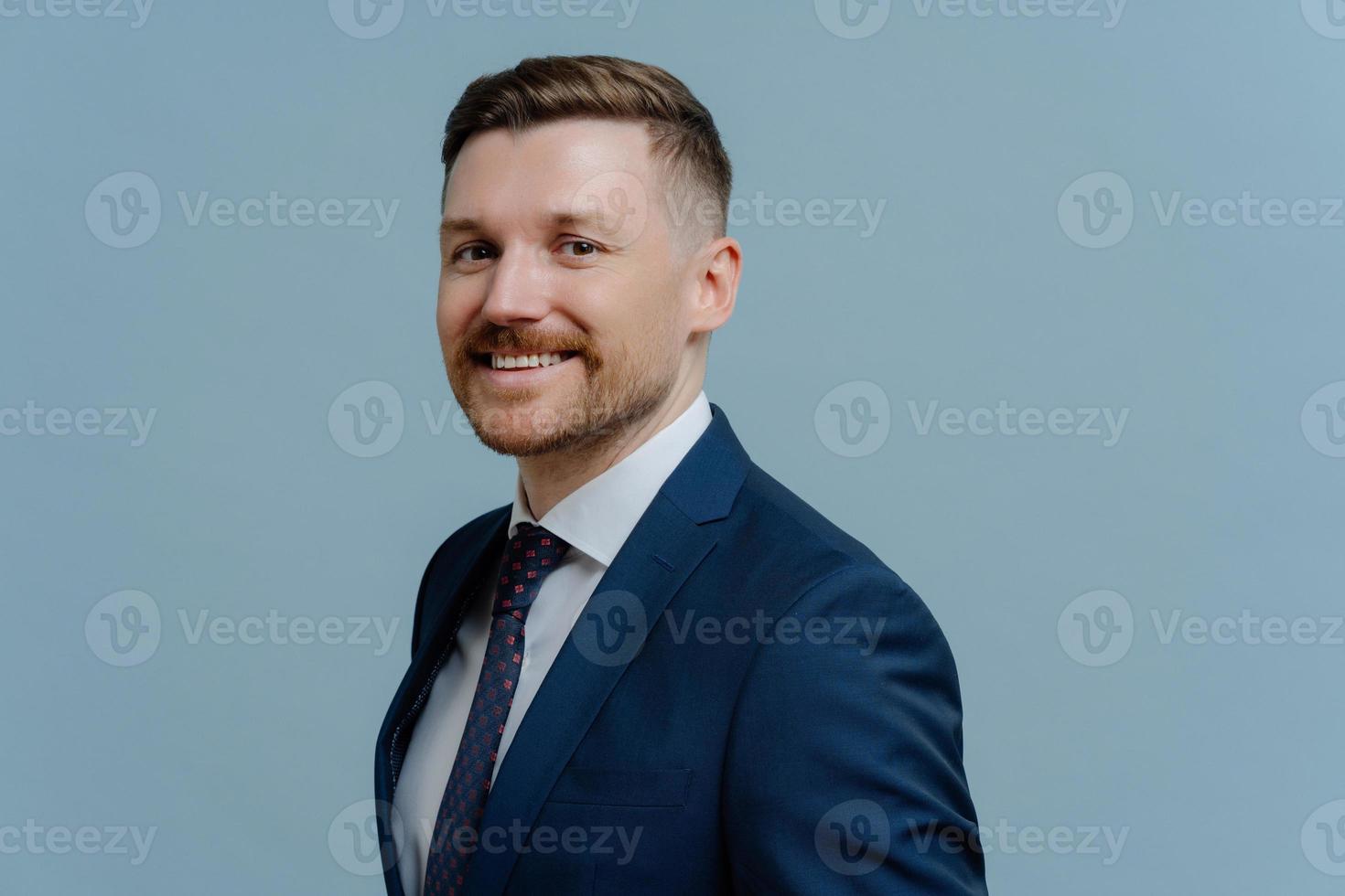 Sideways shot of smiling corporate worker dressed in formal outfit comes on business meeting poses against blue studio background copy space aside. Elegant businessman wears office clothing. photo