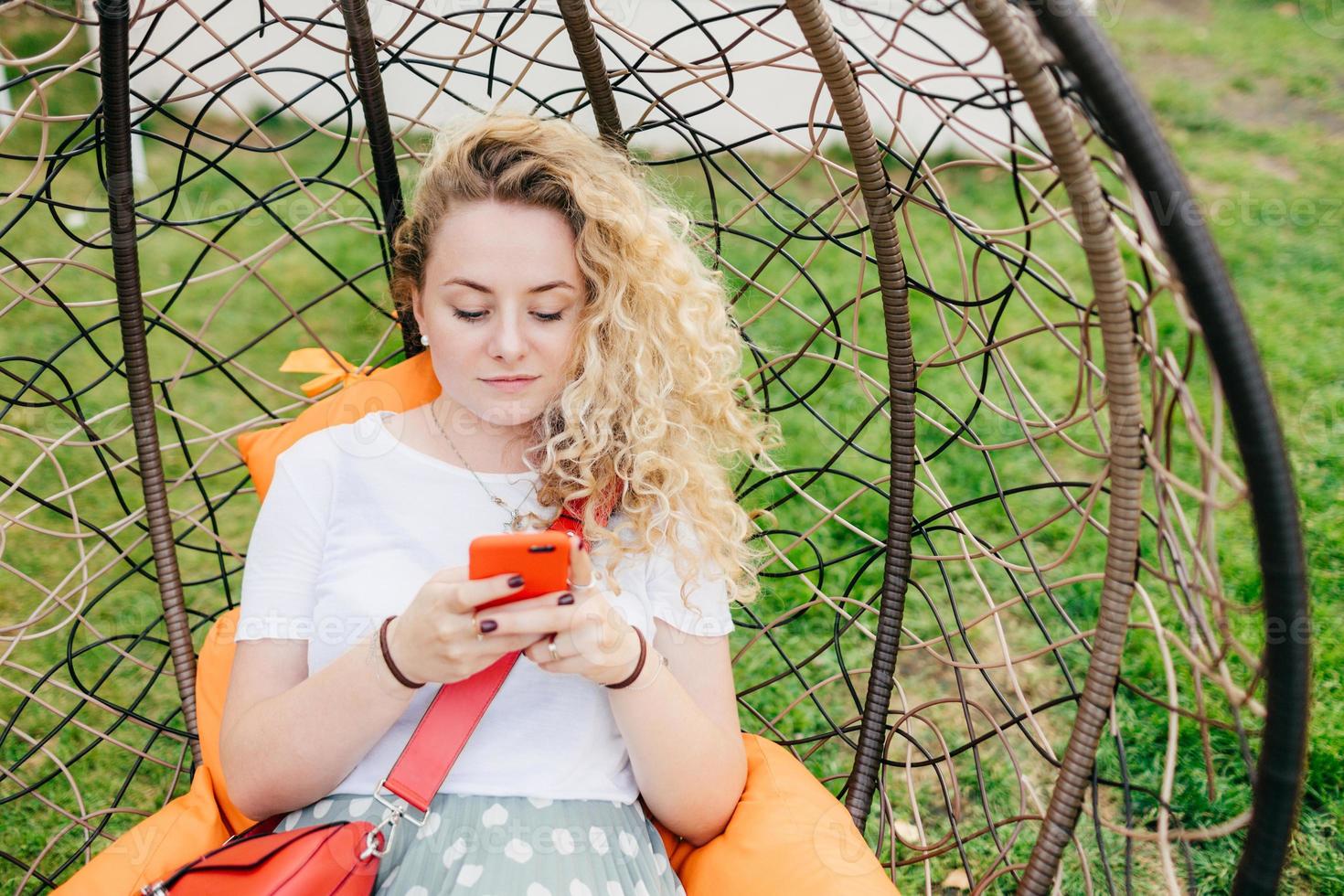 Horizontal shot of pretty young female rests in hanging chair in garden, uses smart phone, being always in touch, checks her email box, dressed casually, has good rest outdoor. Technology concept photo