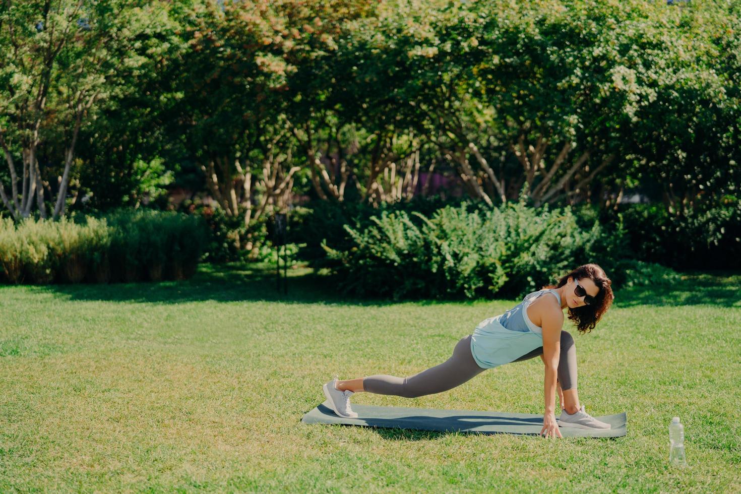 Outdoor sport concept. Healthy fit energetic young woman does fitness exercises on fitness mat dressed in active wear drinks fresh water wears sunglasses poses on green grass. Morning workout. photo