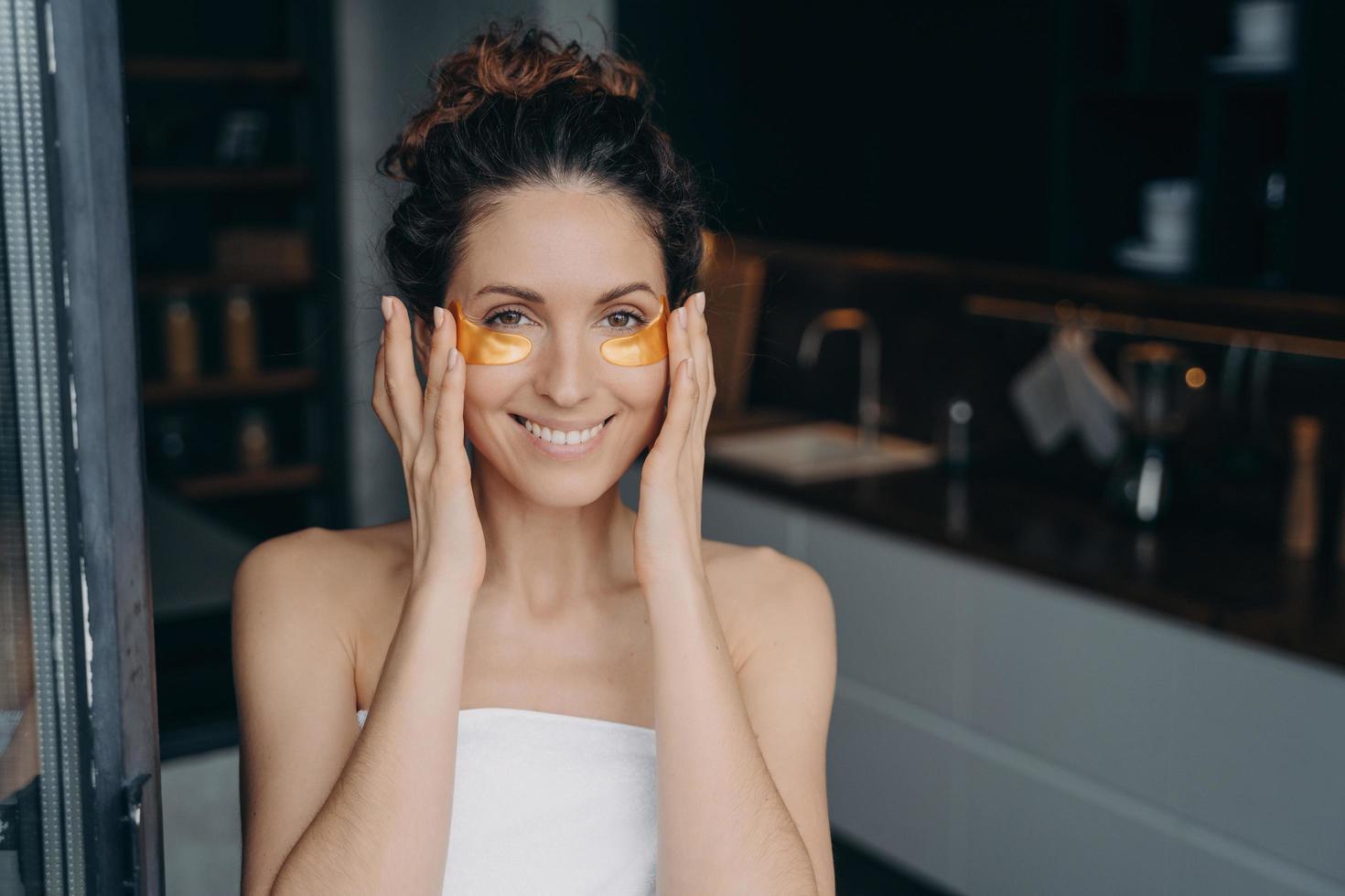 Pretty young lady wrapped in towel after bathing is happy and relaxing applying eye patches. photo