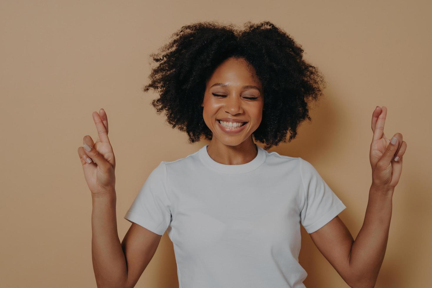 Hopeful dark skinned woman with closed eyes wishing for luck isolated on beige studio background photo