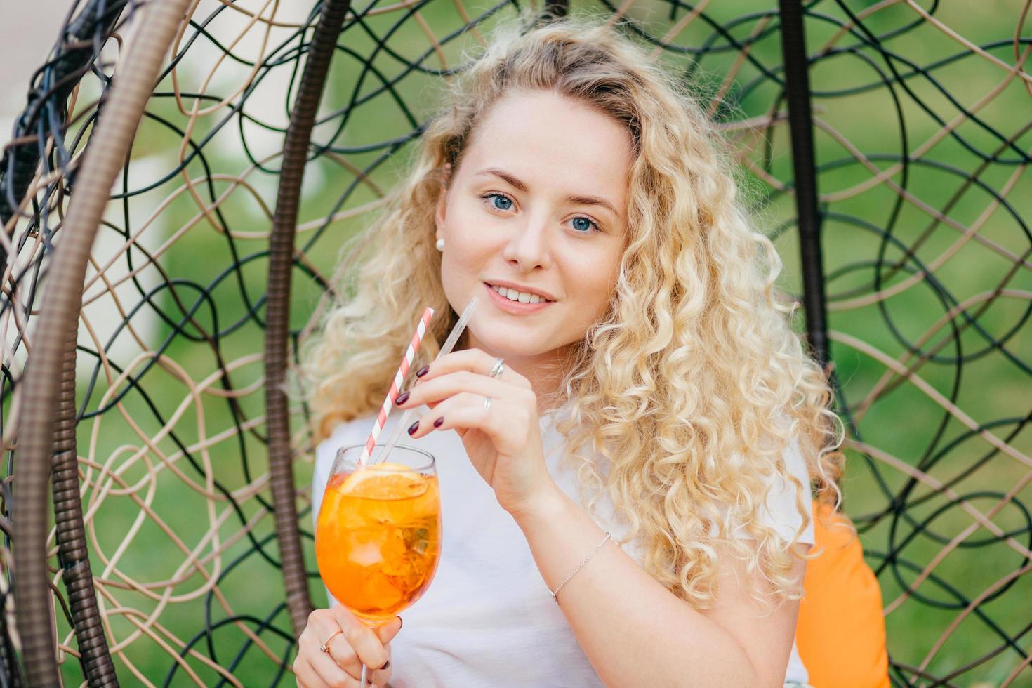 Outdoor shot of pleasant looking female with curly light hair, drinks fresh cold drink, rests in hanging chair, breathes fresh air, has summer party with friends. People and lifestyle concept. photo