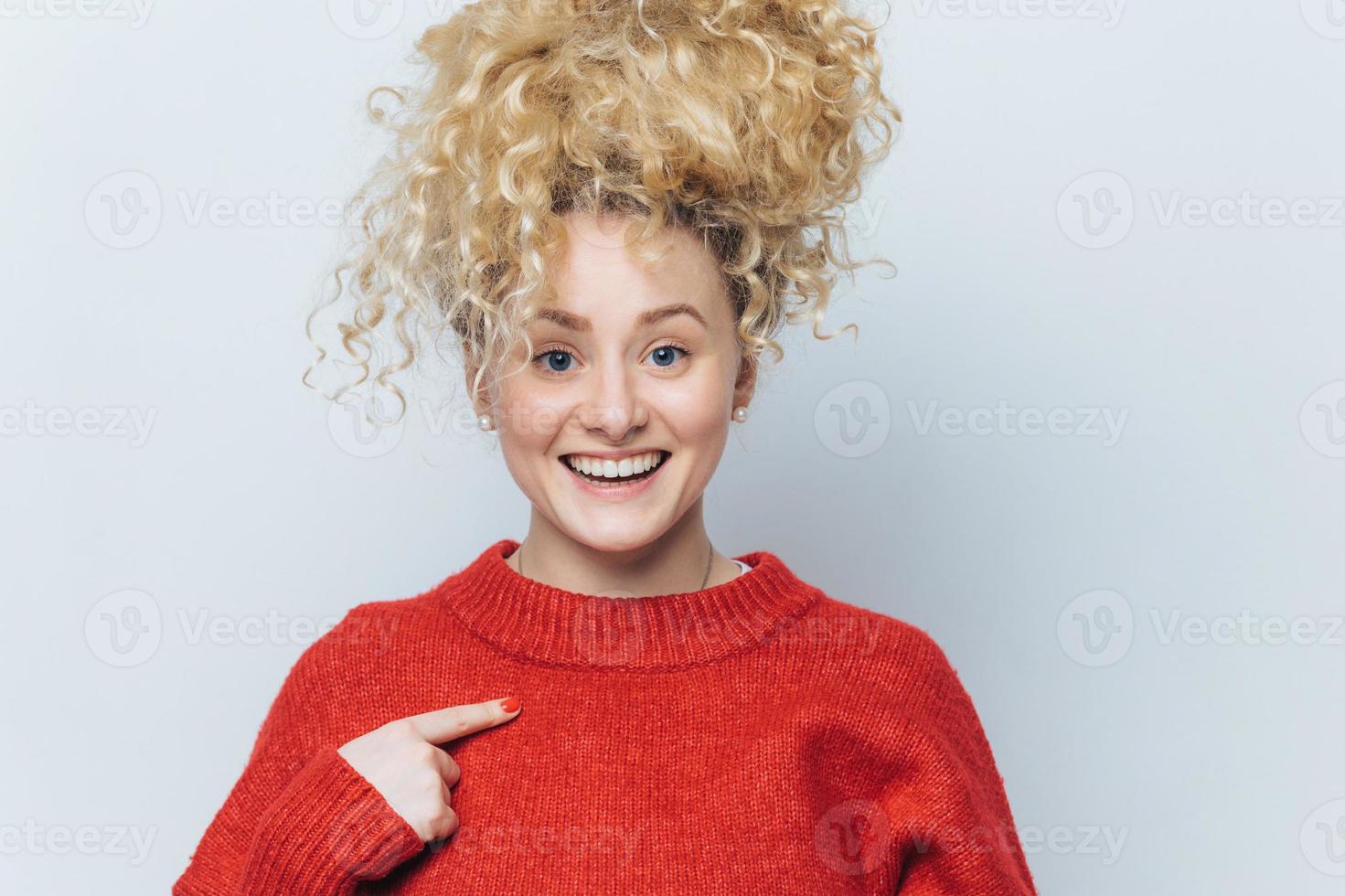 Happy funny beautiful young woman with pleased expression, rejoices her new purchase, indicates at red sweater, shows copy space for your advertisment content. People, style and clothing concept photo