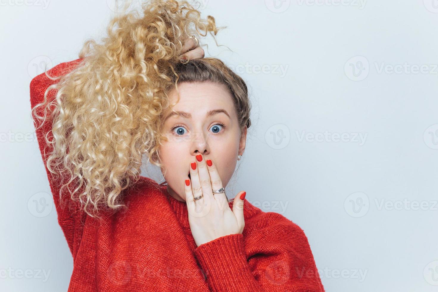Bugged eyed emotional funny blonde female with pony tail and curly hair, covers mouth with hand, doesnt expect to hear shocking news from friend, isolated over white background. Omg concept photo