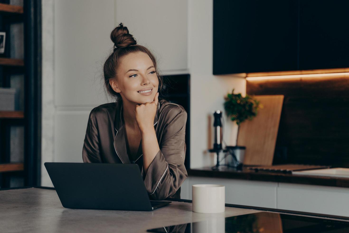Young happy freelancer lady in cozy pajamas working from home on laptop photo