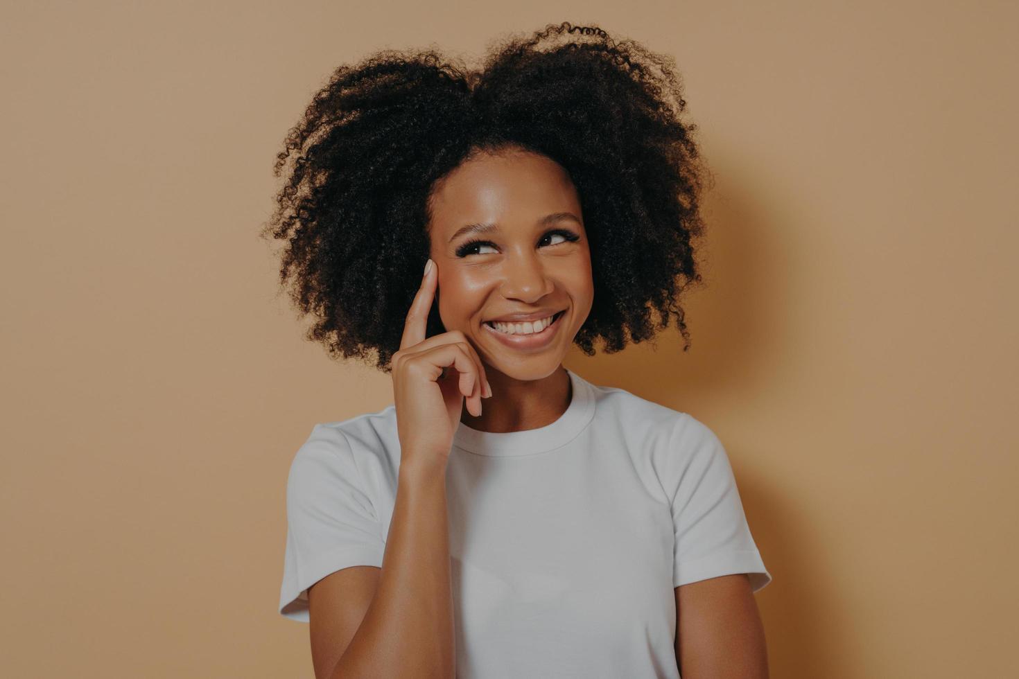 Pensive african woman with curly hair touching temple with forefinger, isolated on beige background photo