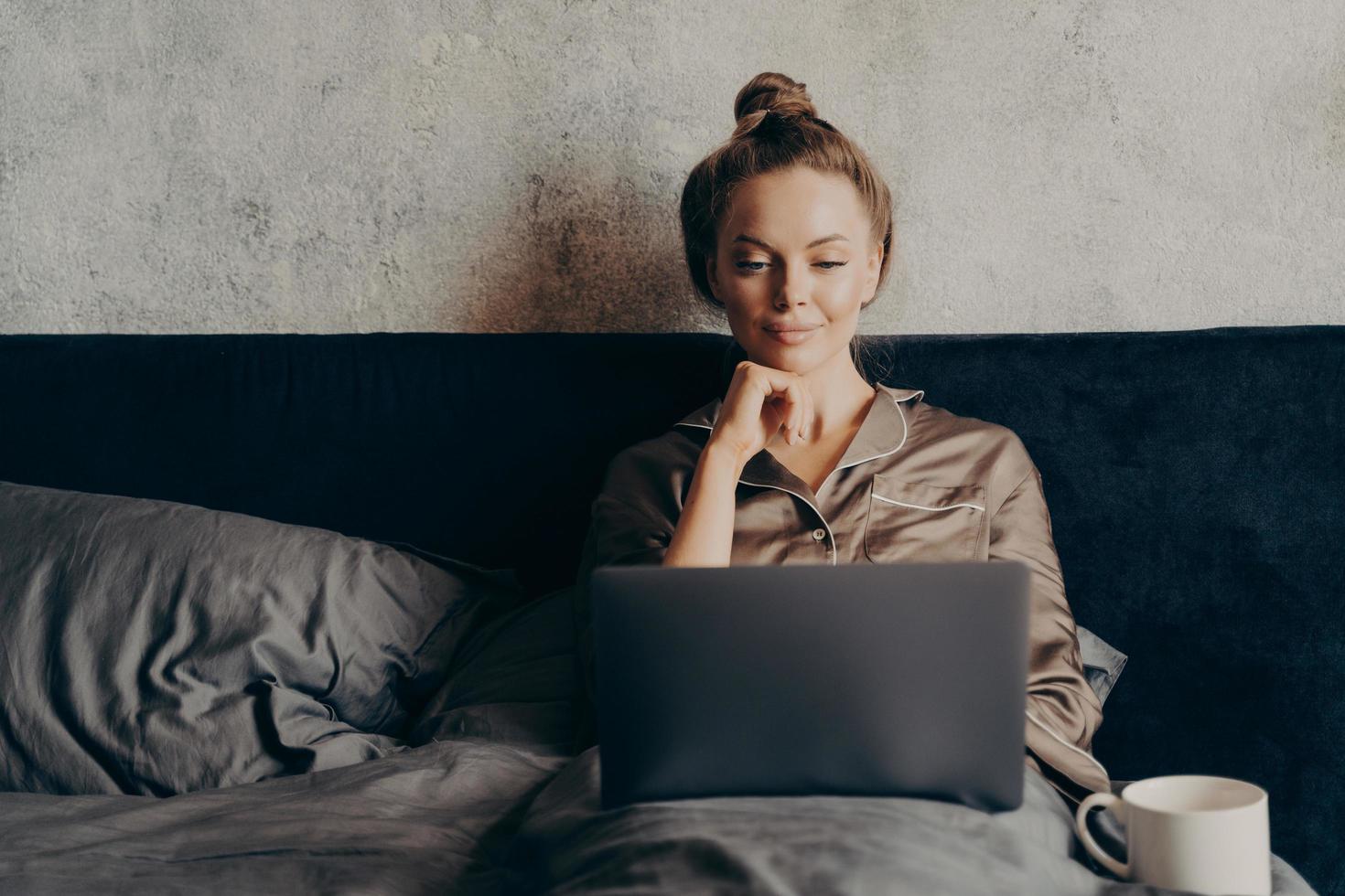 Positive relaxed young woman in pajama lying in bed and using laptop photo