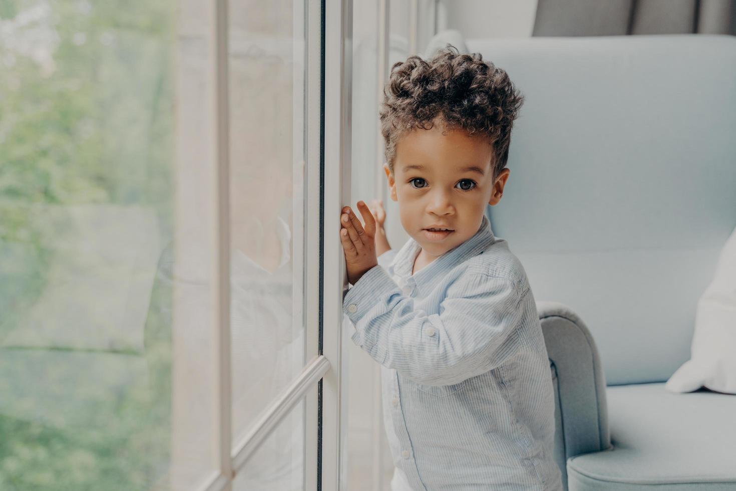 Portrait of cute mixed race curly haired baby boy waiting for parents near window photo