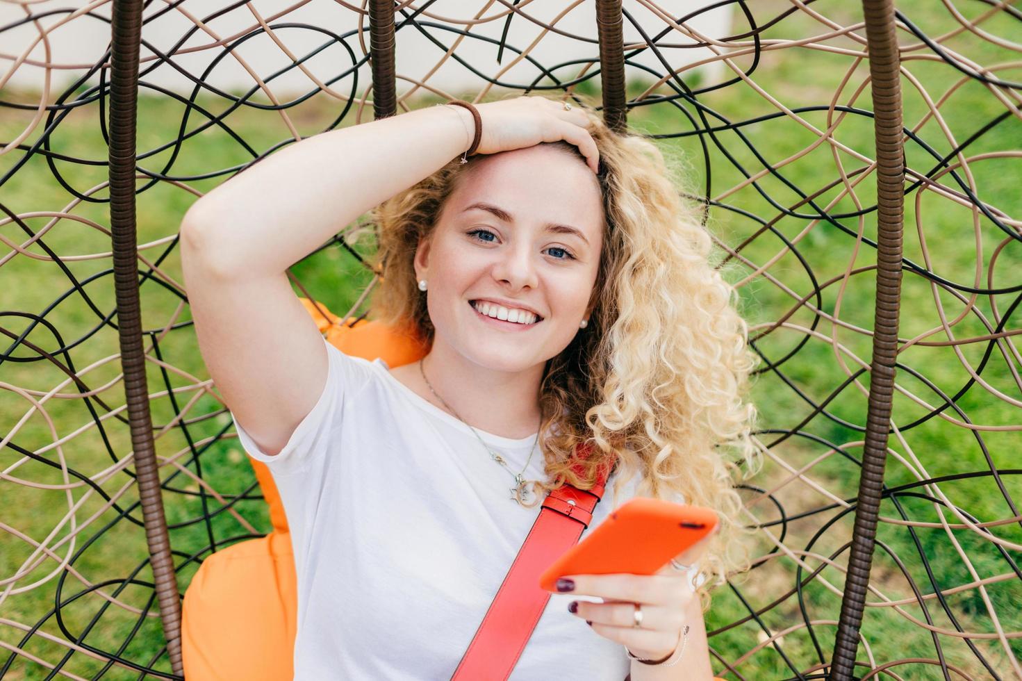 Smiling European female with broad smile, curly hair, dressed in casual white t shirt, uses modern electronic device for surfing social networks, happy to recieve message on invitation to party photo