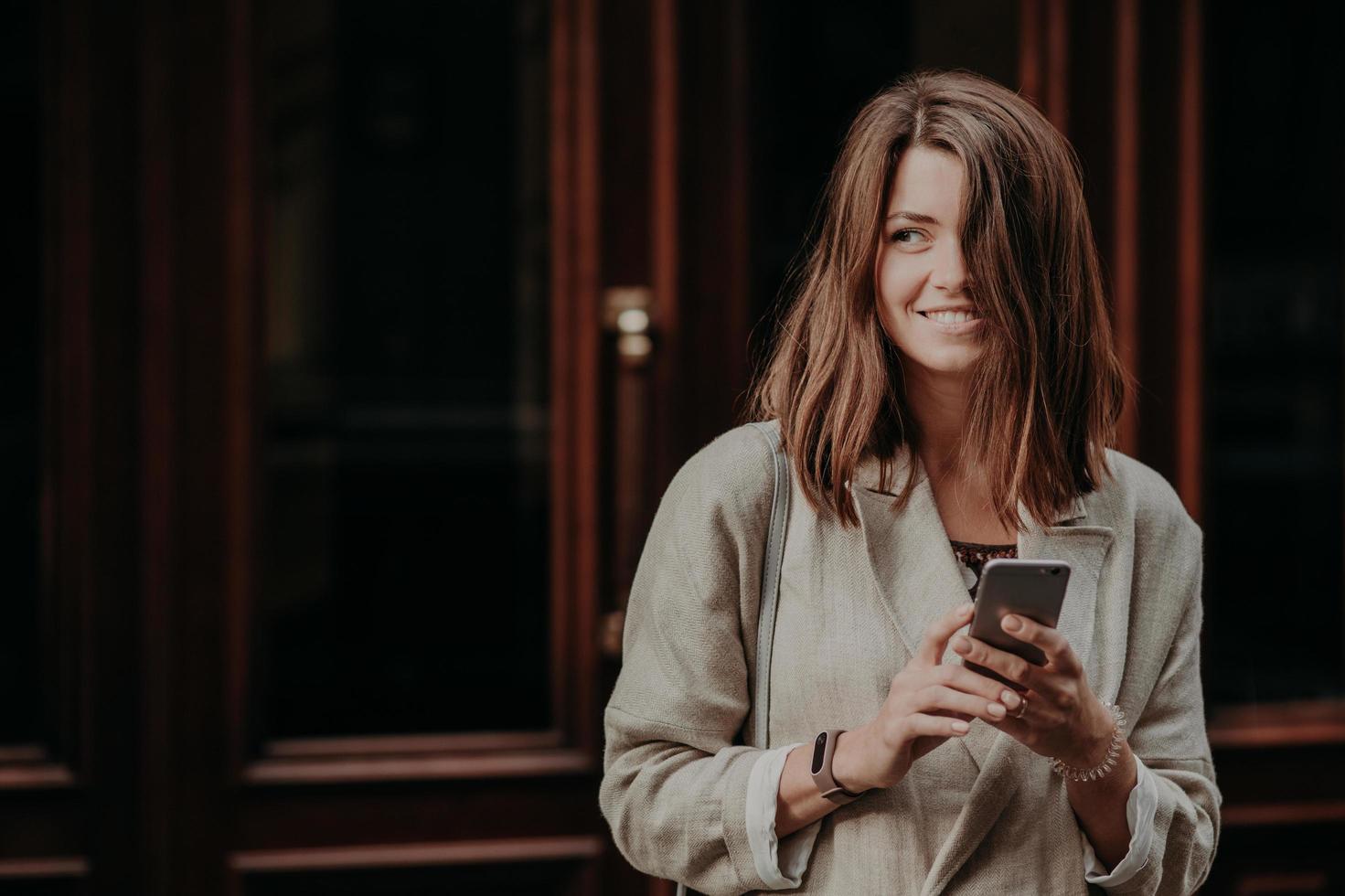 Emotional delighted happy brunette young woman with dark hair, uses mobile phone for texting messages, dressed in elegant raincoat, stands near door, cant imagine life without modern technoliges photo
