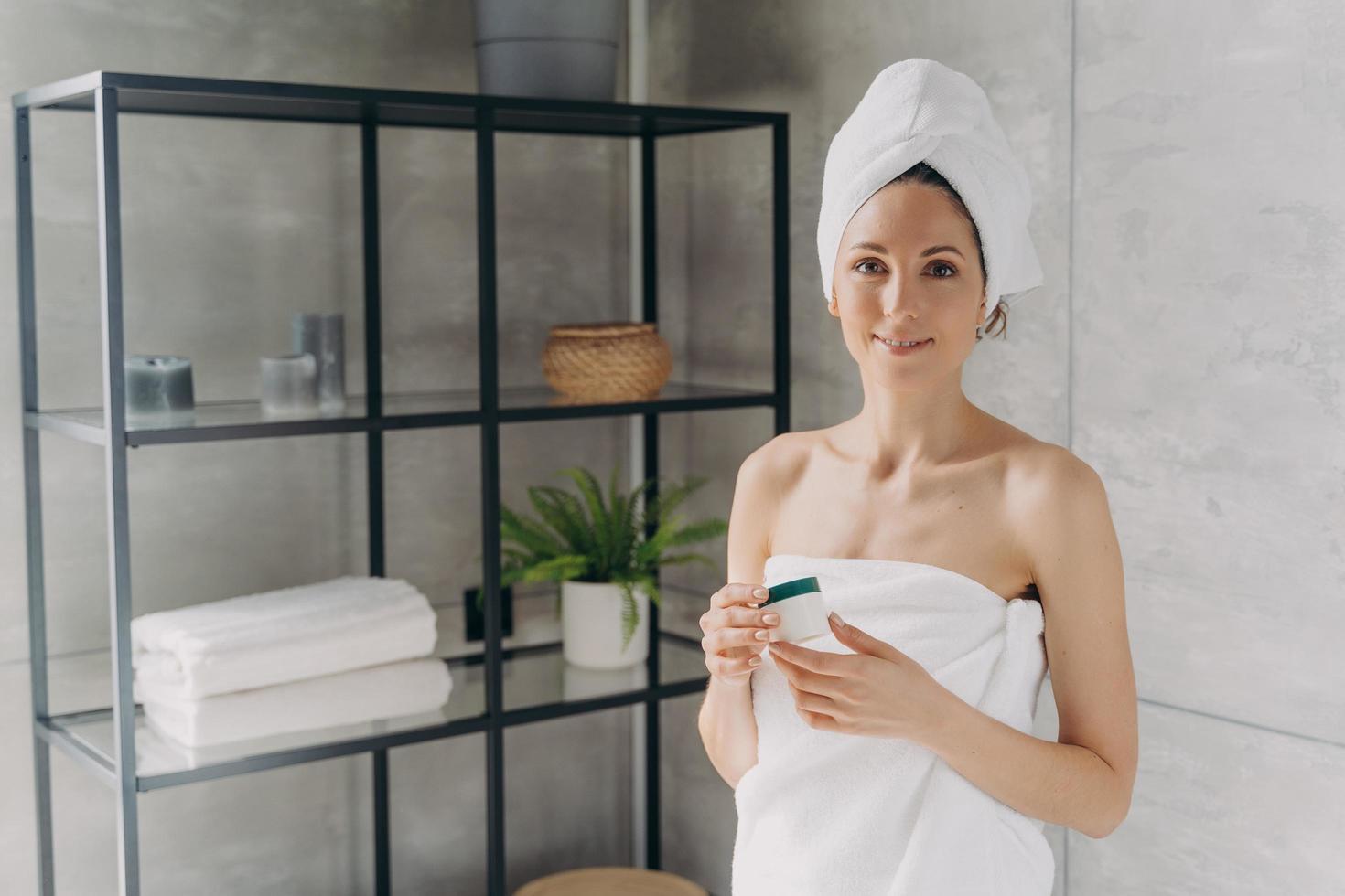 Attractive caucasian girl wrapped in towel after bathing and hair washing is holding a cream jar. photo