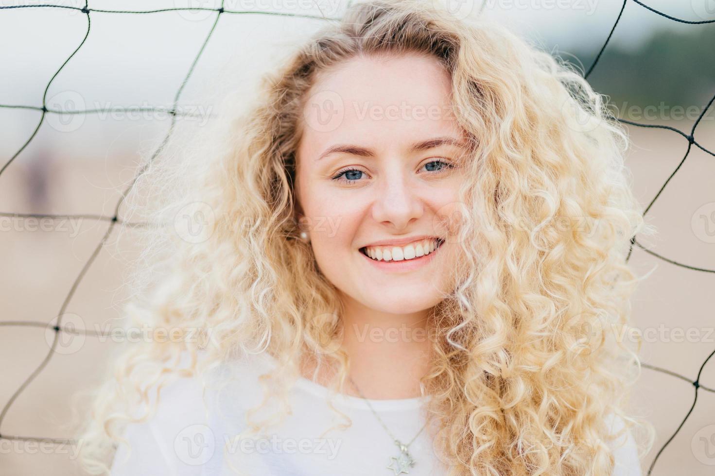 Lovely cute young woman has fresh skin, curly light hair, smiles gladfully, dressed in casual clothes, stands near tennis net, being satisfied to recieve compliment. People, happiness concept photo