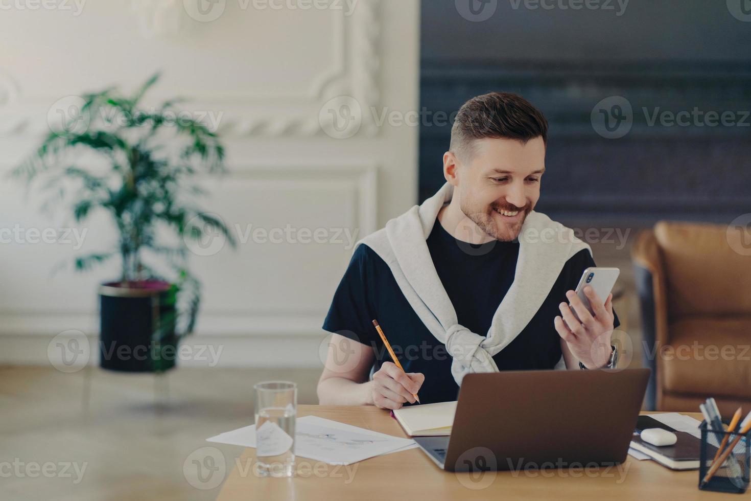 Happy bearded young man studies indoor with modern gadgets writes down notes from internet website makes records in notebook holds smartphone poses at coworking space cozy interior. Hipster student photo