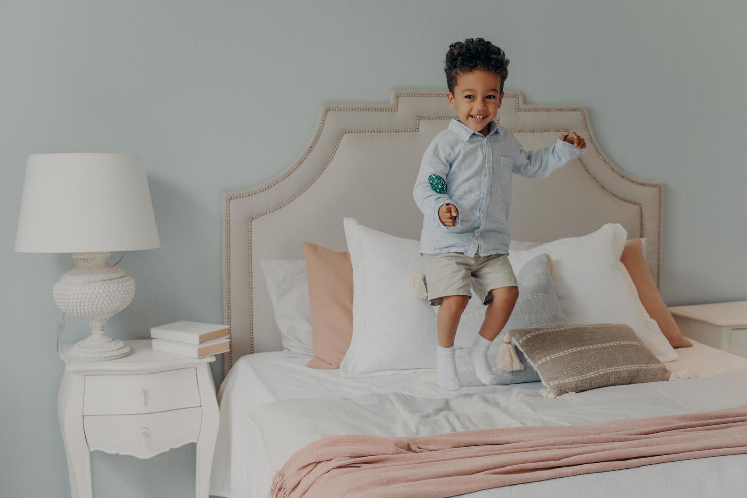 niño afroamericano enérgico saltando con piruleta en el colchón de la cama foto