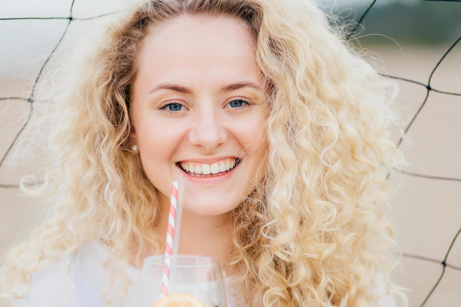 Close up portrait of cheerful young female with curly hair, smiles positively, holds cold drink in front, stands against net background, being in good mood after date with boyfriend. Beauty concept photo