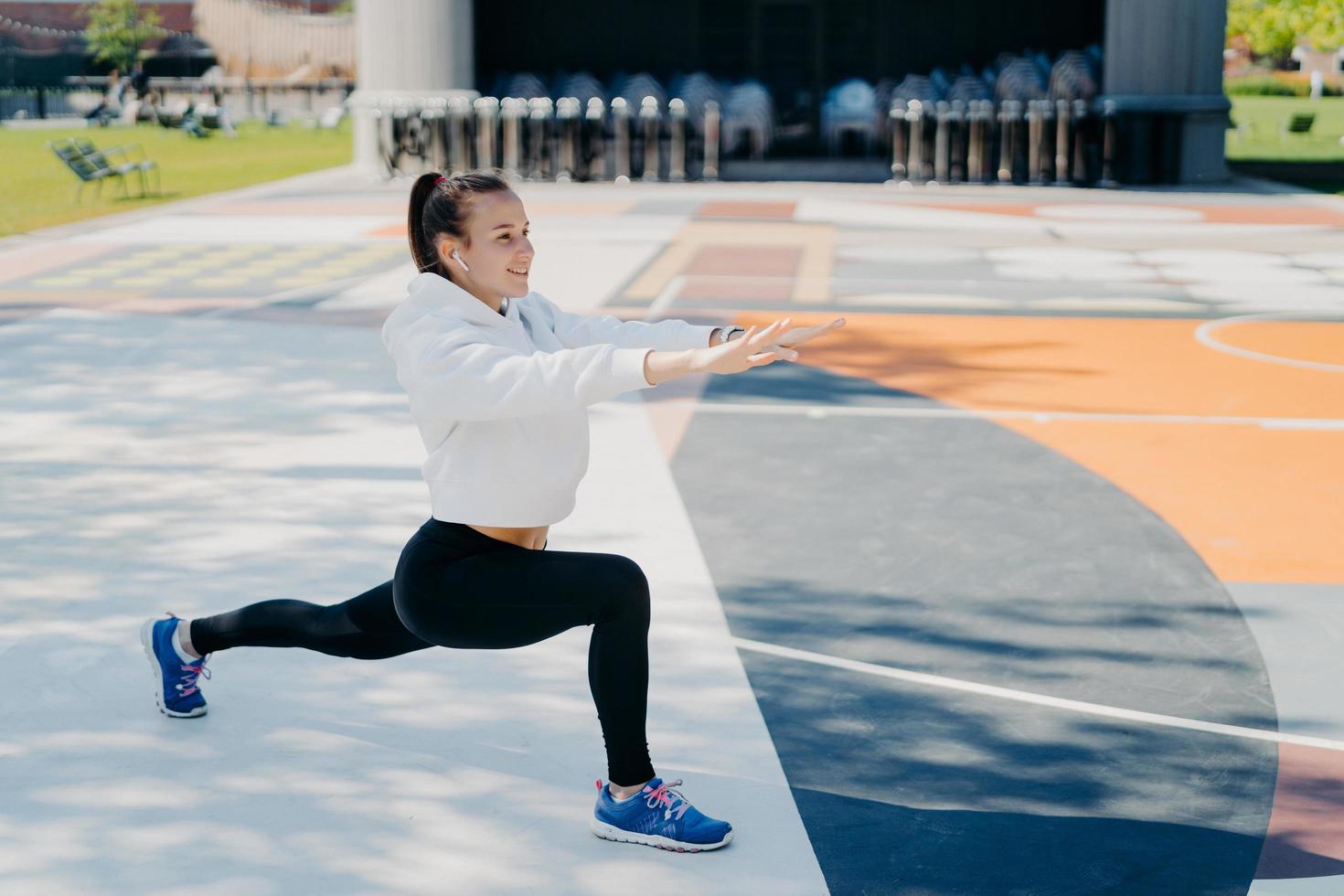 Horizontal shot of motivated sporty woman stretches legs and arms does warming up exercises before workout looks forward dressed in sportsclothes poses outside on big stadium. Exercising concept photo