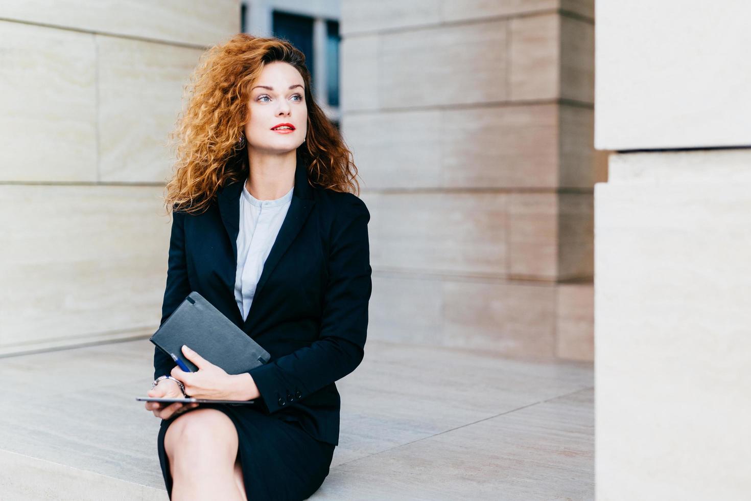 Good-looking young businesswoman with luxuriant hair, blue eyes, red lips, wearing black suit, holding in hands pocket book and modern tablet computer, looking aside with thoughtful expression photo