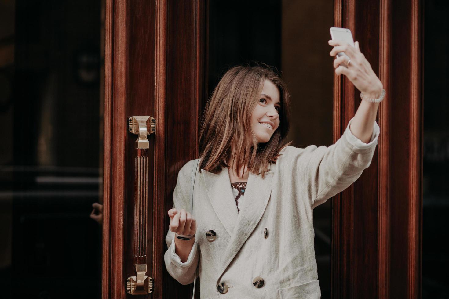 Pretty adorable woman makes selfie with smart phone, dressed in raincoat, poses outdoor, uses modern technology, shares photos in social networks. People, technology and entertainment concept