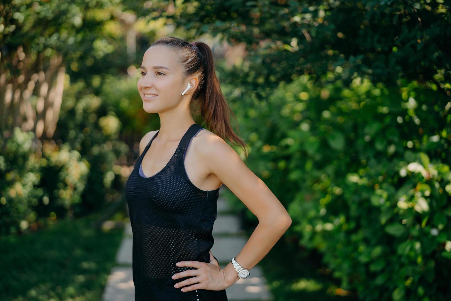 Half length shot of sporty young woman dressed in sportswear keeps hands on waist has dark hair combed in pony tail focused into distance with happy smile on face listens music poses outdoors photo