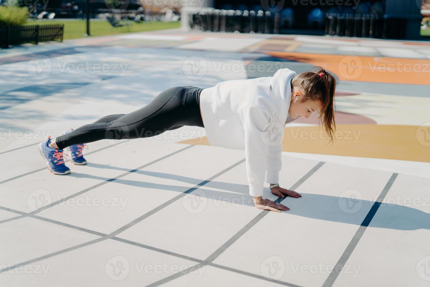 Full length shot of active young woman with pony tail stands in plank focused down dressed in active wear poses outdoors being motivated gets stronger demonstrated endurance and determination photo