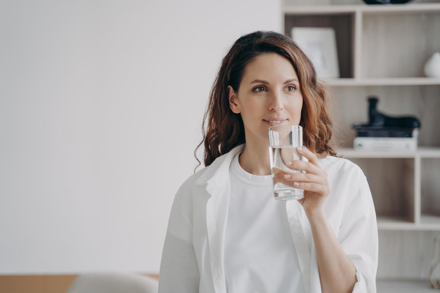 Relaxed european woman is drinking pure water at home. Happy girl is holding glass and smiling. photo