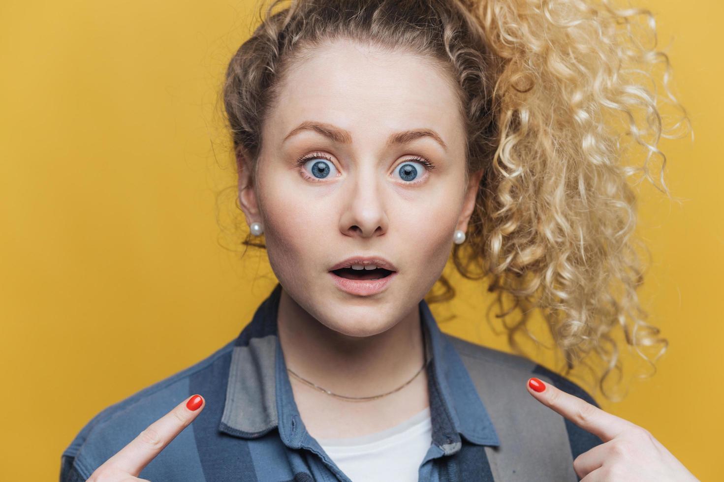 Horizontal shot of pleasant looking young curly woman indicates with fore fingers at herself, being shocked to recieve failure, dressed casually, has amazed expression, isolated over yellow wall photo