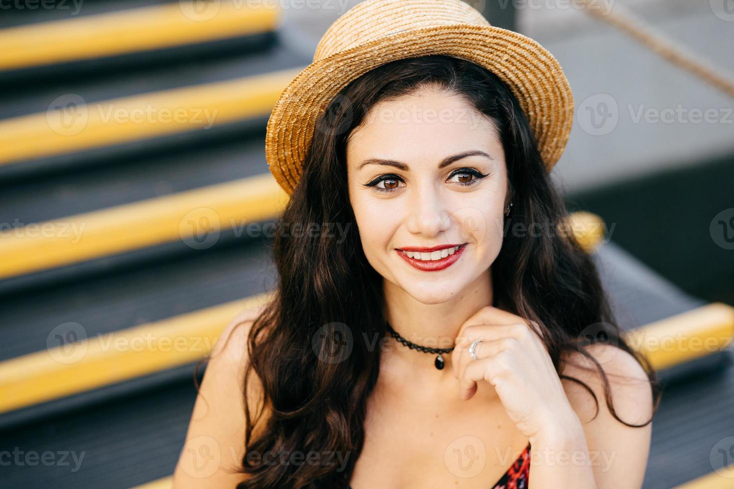 Outdoor portrait of fashionable woman with dark thick hair having make-up and painted red lips wearing summer straw hat and necklace looking into distance having gentle smile sitting at stairs photo