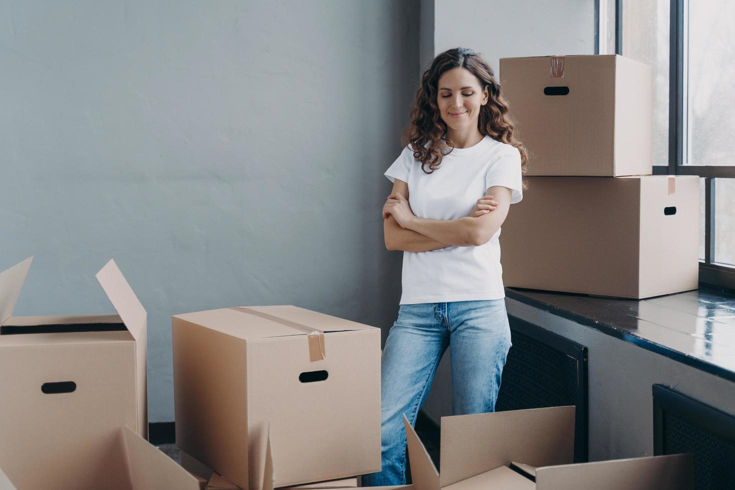 Satisfied woman in jeans and white t-shirt unpacking boxes. Attractive girl in new apartment. photo