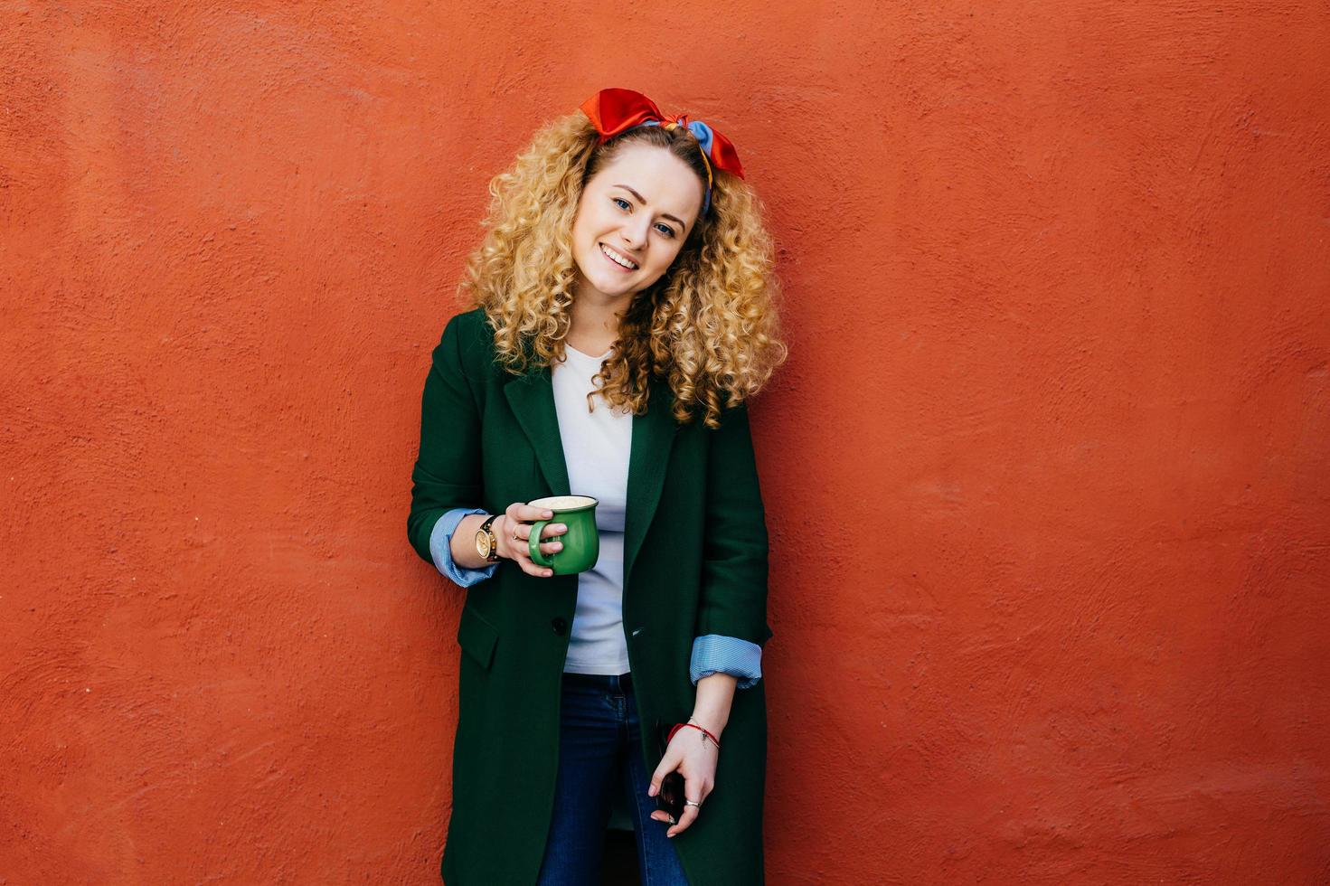 Young fashionable Caucasian woman with curly hair wearing headband and stylish jacket holding green cup of coffee having happy expression while resting against orange background. Woman with coffee mug photo