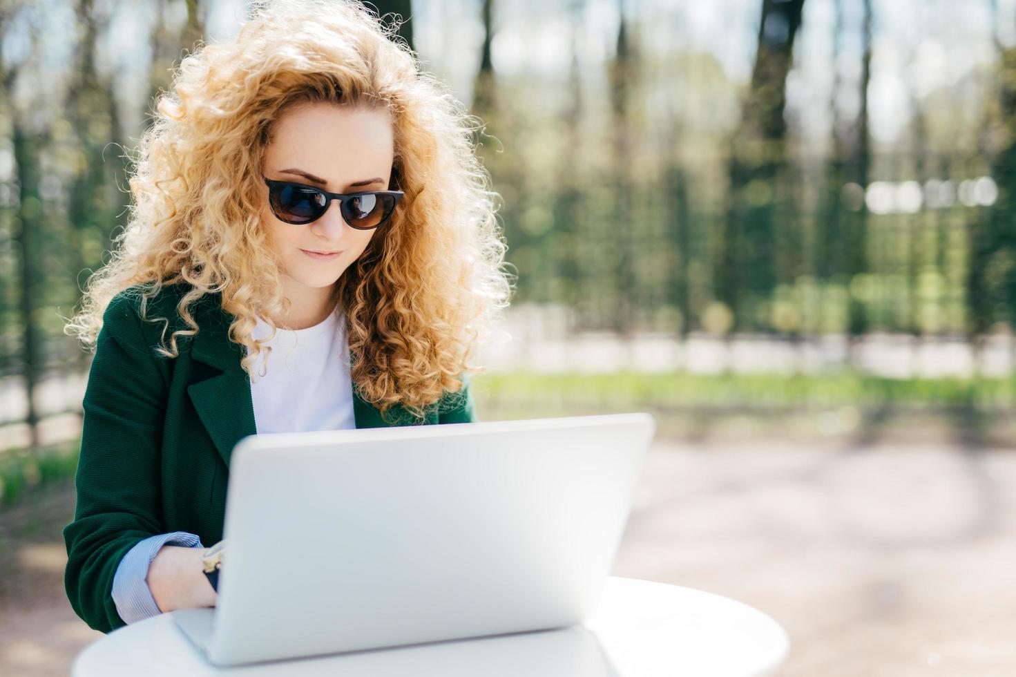 Serious female student having fluffy blonde hair wearing green jacket and sunglasses sitting outdoors making her diploma paper enjoing beautiful sunny weather. Fashion, beauty, youth concept photo