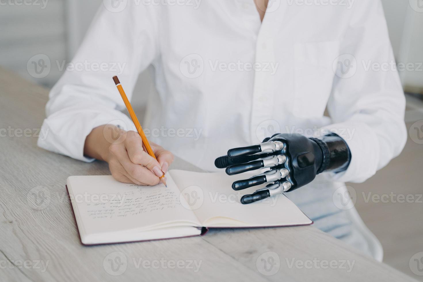 Hands of woman which is taking notes with pencil. Cyber arm and healthy limb on the notebook. photo